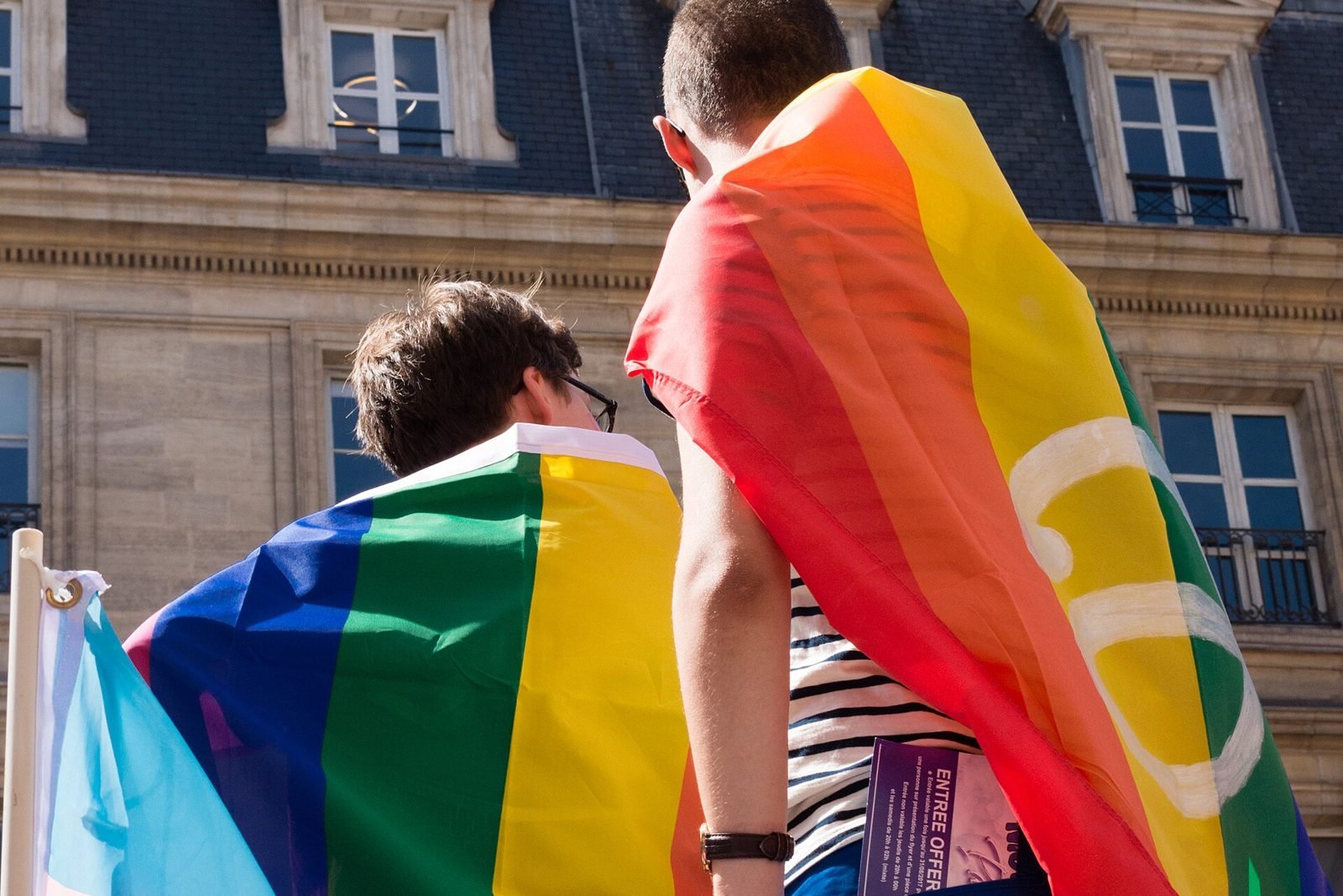 Deux jeunes adultes de dos, drapeaux arc-en-ciel sur les épaules, lors de la Marche des fiertés à Paris